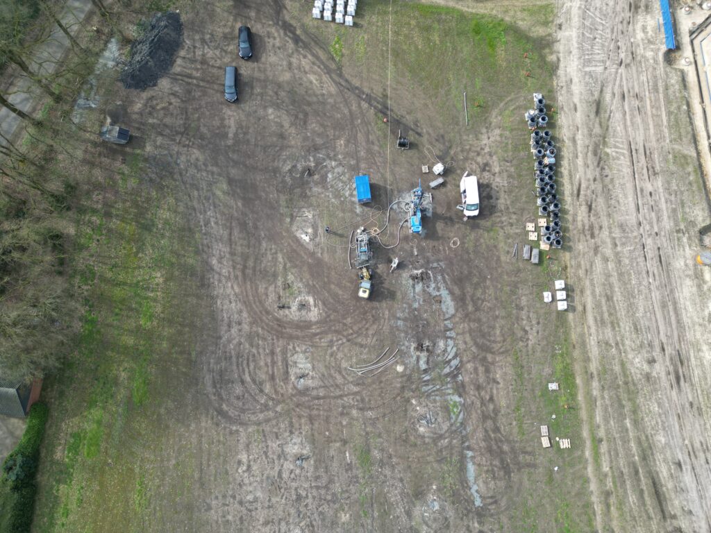 Drohnenaufnahme einer Baustelle auf freiem Feld mit Baugerät, Kabeltrommeln, mehreren Fahrzeugen und Stapeln von Rohren rechts.