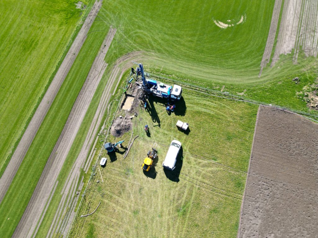 Bau- und Landmaschinen auf einer Wiese: Bohrgerät an Lkw, Traktor, Transporter und Arbeitsbereich mit Fahrspuren und grünen Feldern aus der Vogelperspektive.