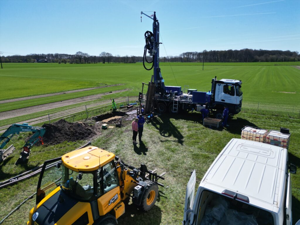 Bohrgerät auf Laster und Bagger auf einer Baustelle am Feldrand, Arbeiter neben offenem Schacht bei grünem Ackerland und blauem Himmel