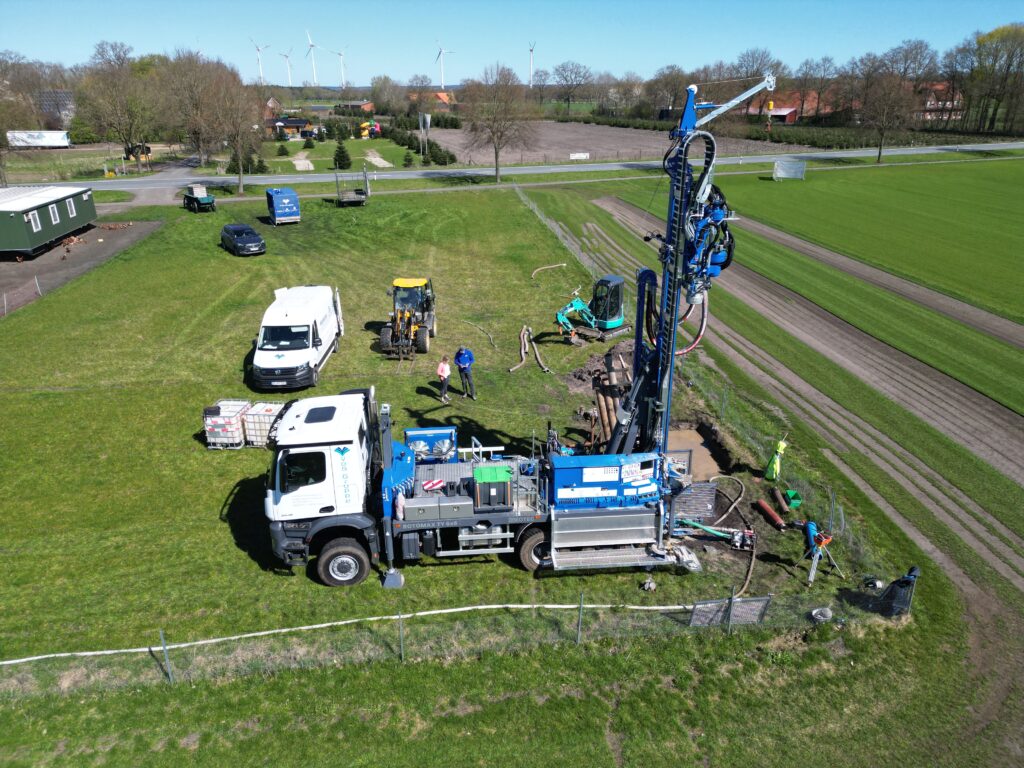 Bohrtruck mit ausgefahrener Bohrmast auf einer Wiese, daneben Transporter, Bagger und zwei Personen, Windräder im Hintergrund
