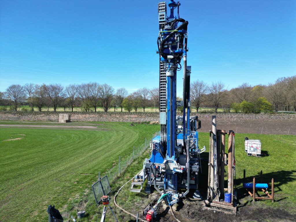 Große blaue Bohrmaschine auf einem Feldrand, Maschinenmast mit Schläuchen und Hydraulik, Holzpfähle und Paletten im Hintergrund