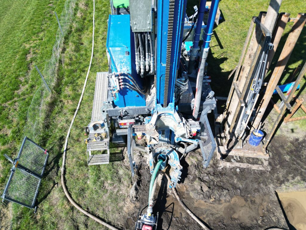 Blick von oben auf blaues Bohrgerät mit Schläuchen, Metallrohren und Pfahlgestell auf einer grasigen Baustelle neben schlammiger Baugrube